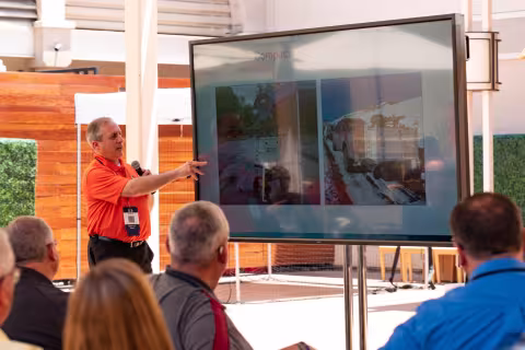 A FirstNet Authority 5x5 presenter holding a microphone gestures to a large screen television with images of FirstNet Compact Rapid Deployable assets as a seated audience observes