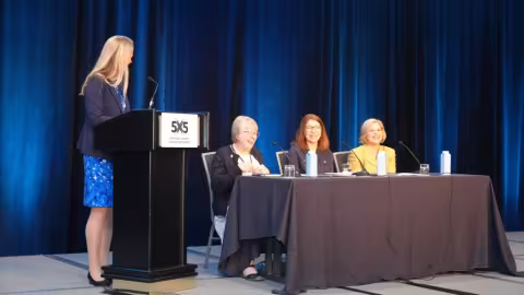 The FirstNet Authority’s Jennifer Harder moderates a panel discussion from behind a podium with a 5x5 placard, three public safety leaders are seated at a table beside the podium