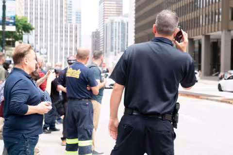 A law enforcement officer stands near a crowd on a sidewalk outside 5x5, speaking into a cell phone